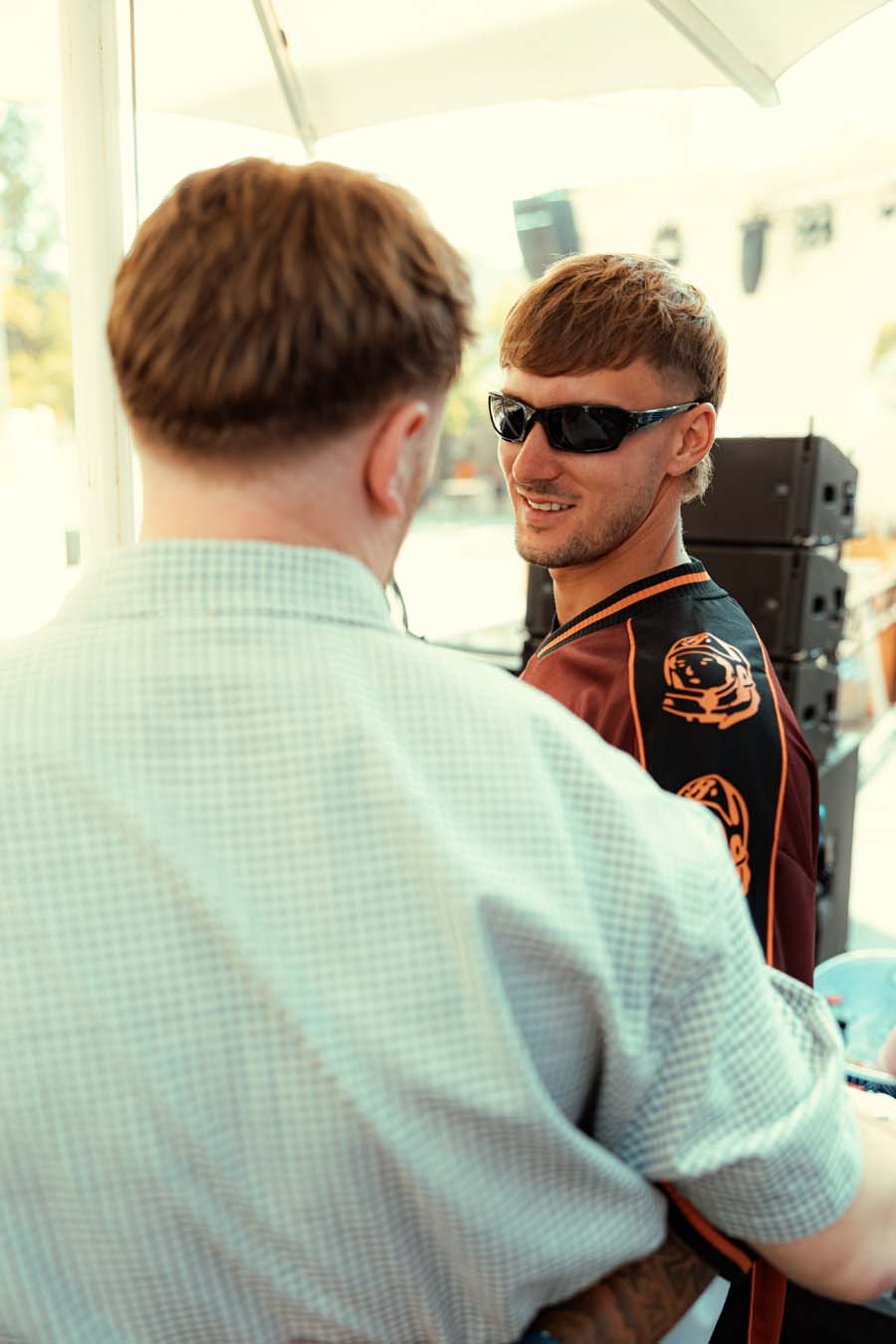 A man in a maroon and black varsity-style jacket with orange trim and a tiger graphic on the sleeve, wearing black sunglasses, is seen from the side while another person in a light green checkered shirt is shown from behind in an outdoor setting under a white canopy.