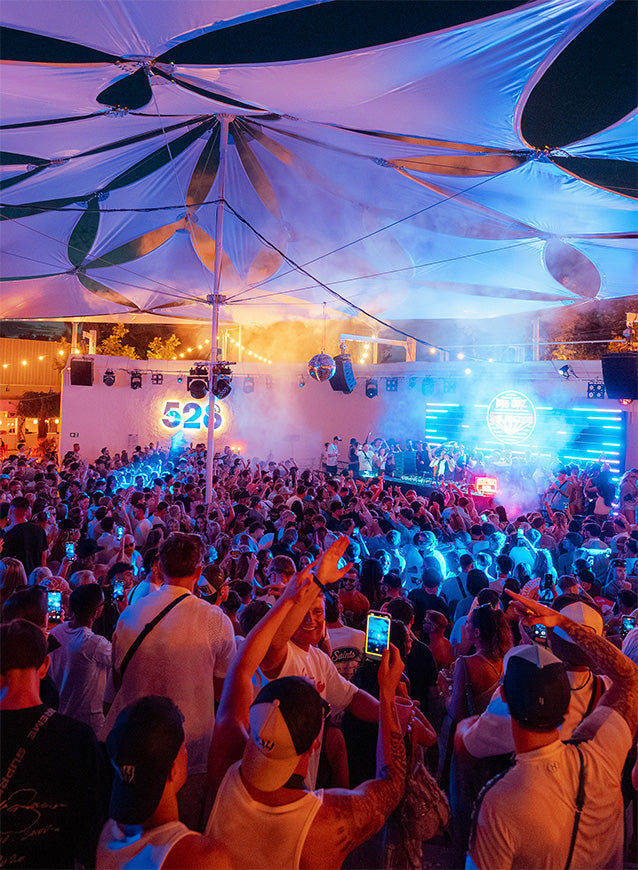 A crowded outdoor night festival under a large white canopy with blue and orange stage lighting, featuring a live performance on a raised platform and many people holding up phones in the foreground.