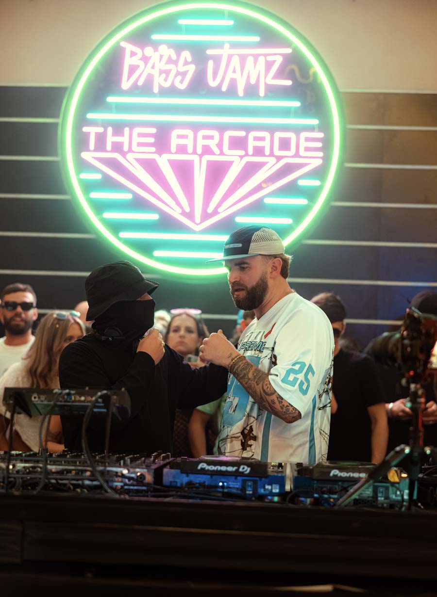 A man in a white graphic t-shirt and cap stands behind DJ equipment at an event, with another person facing him. A large neon sign in the background reads 'Bass Jamz The Arcade' in green and pink.