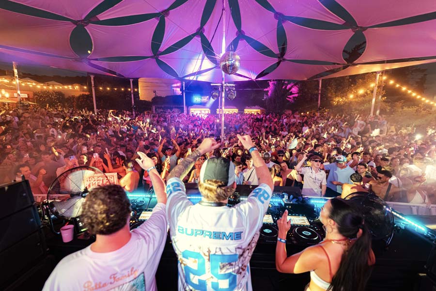 A crowded outdoor concert scene under a large patterned canopy, with DJs performing at turntables in front of a large audience. A person in the center wears a white Supreme jersey with blue lettering and the number 25 on the back.