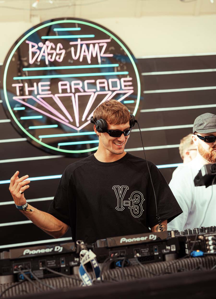 A man wearing a black Y-3 t-shirt and sunglasses stands behind DJ equipment at a live event, with a neon sign reading 'Bass Jamz The Arcade' in the background.