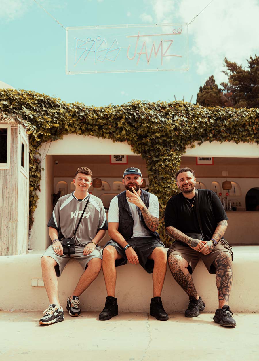 Three men sitting together outdoors in front of a building with ivy-covered walls and a blue sky above. A neon-style sign at the top reads 'Bass Jamz'.