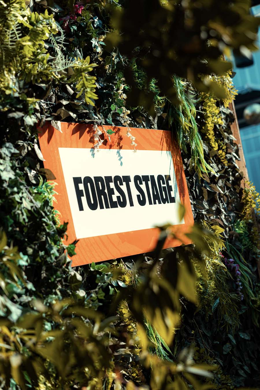 A rectangular orange and white sign reading 'FOREST STAGE' in bold black letters, attached to a leafy green wall and partially framed by blurred foliage in the foreground.