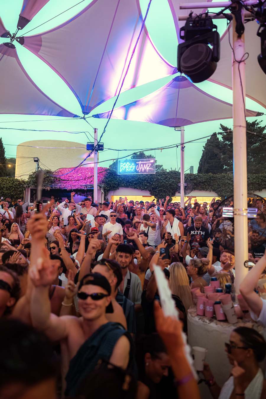 A crowded outdoor night event under large white canopy tents with colorful LED lighting in purple, blue, and pink. A large audience of people is dancing and raising their hands, with stage lighting and speakers visible.