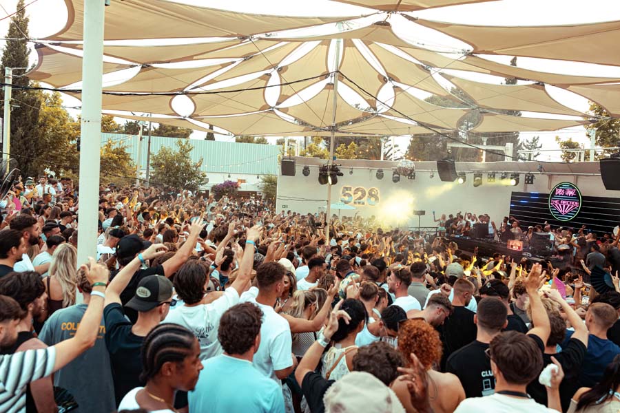 A large crowd of people dancing and raising their hands under a beige canopy tent at an outdoor music festival, with a stage and stage lights visible in the background.