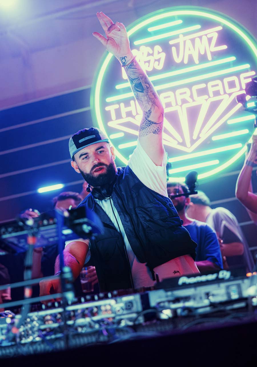 A bearded man with a tattooed arm wearing a black vest and a black cap, posing with his hand raised in front of a glowing green neon sign that reads 'Jazz Jamz The Arcade'. He is standing behind DJ equipment at a club.