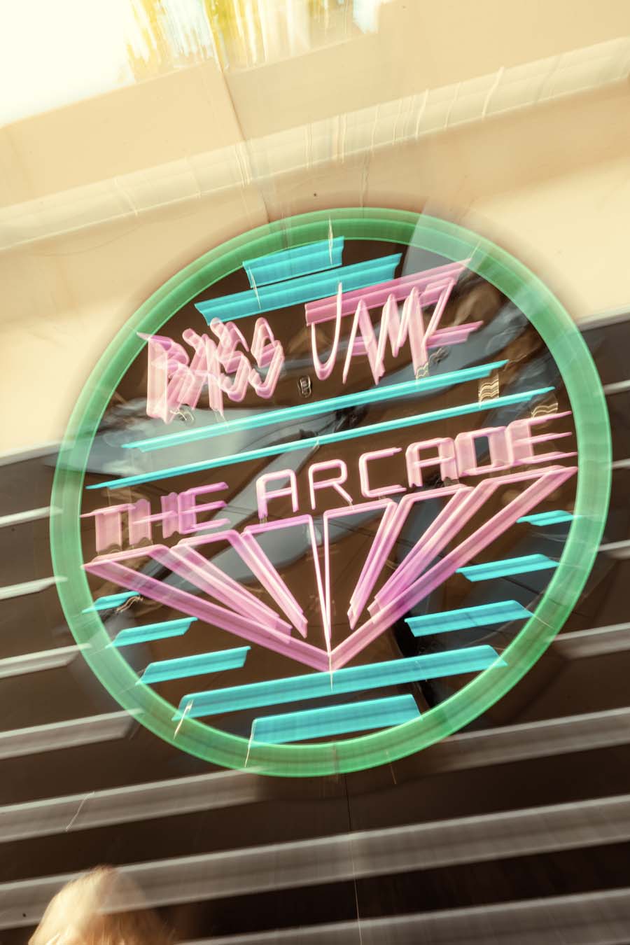 A circular neon sign for 'Bass Jamz The Arcade' glowing in pink and teal against a dark background, mounted on a building exterior.