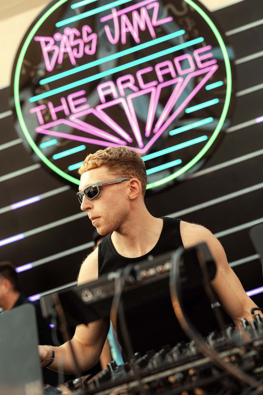 A man wearing sunglasses and a black tank top stands behind DJ equipment in a club setting, with a large neon sign reading 'Bass Jamz The Arcade' in the background.