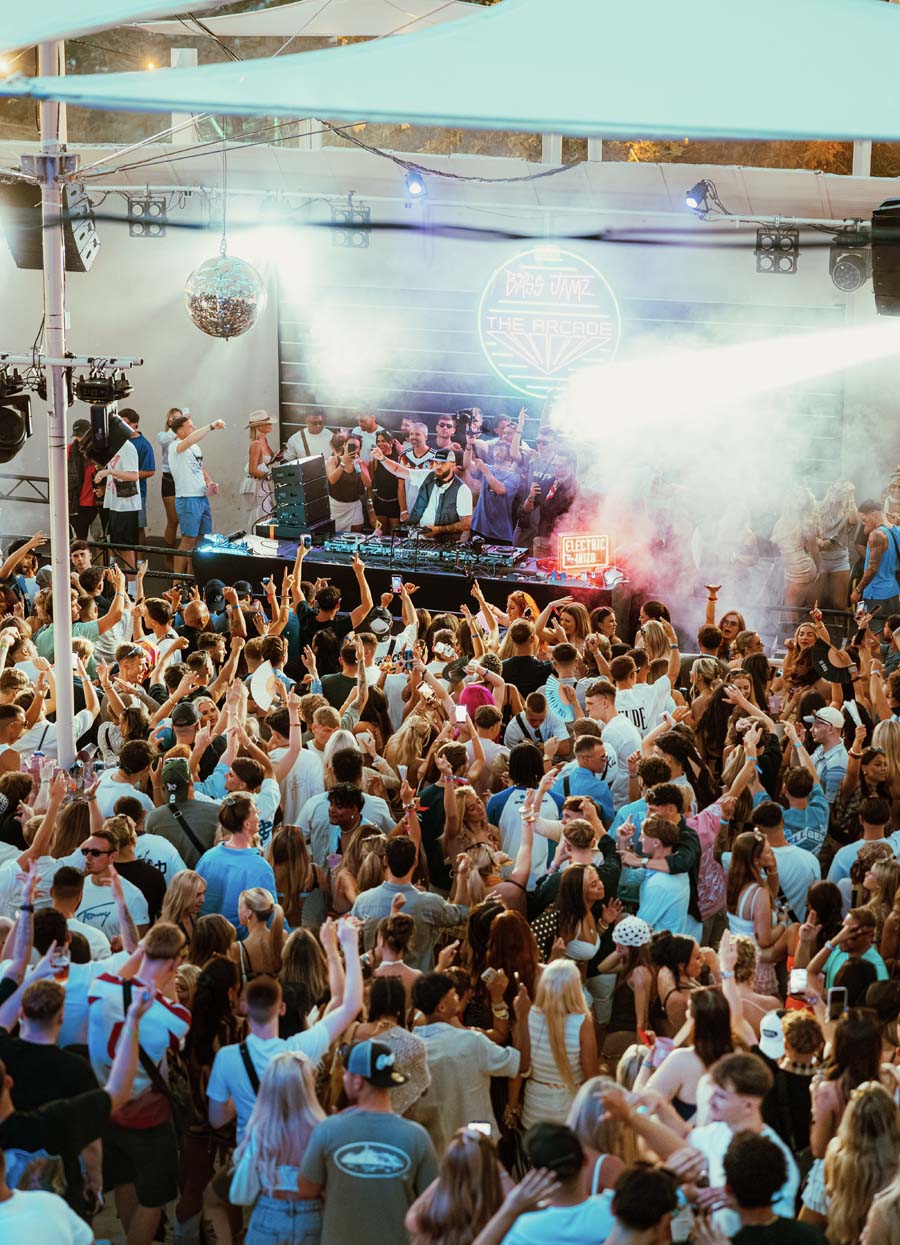 A crowded outdoor concert or festival scene with a large audience facing a stage where a performer is on a lit-up stage under a canopy. Smoke, stage lights, and a disco ball are visible, with many people raising their hands.
