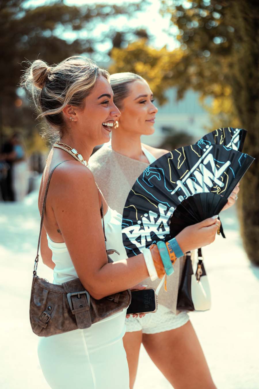 Two women walking outdoors in a sunny, warm-toned setting. The woman in the foreground is wearing a white dress and carrying a brown crossbody bag, while the woman behind her holds a black folding fan with blue and white graphic text.