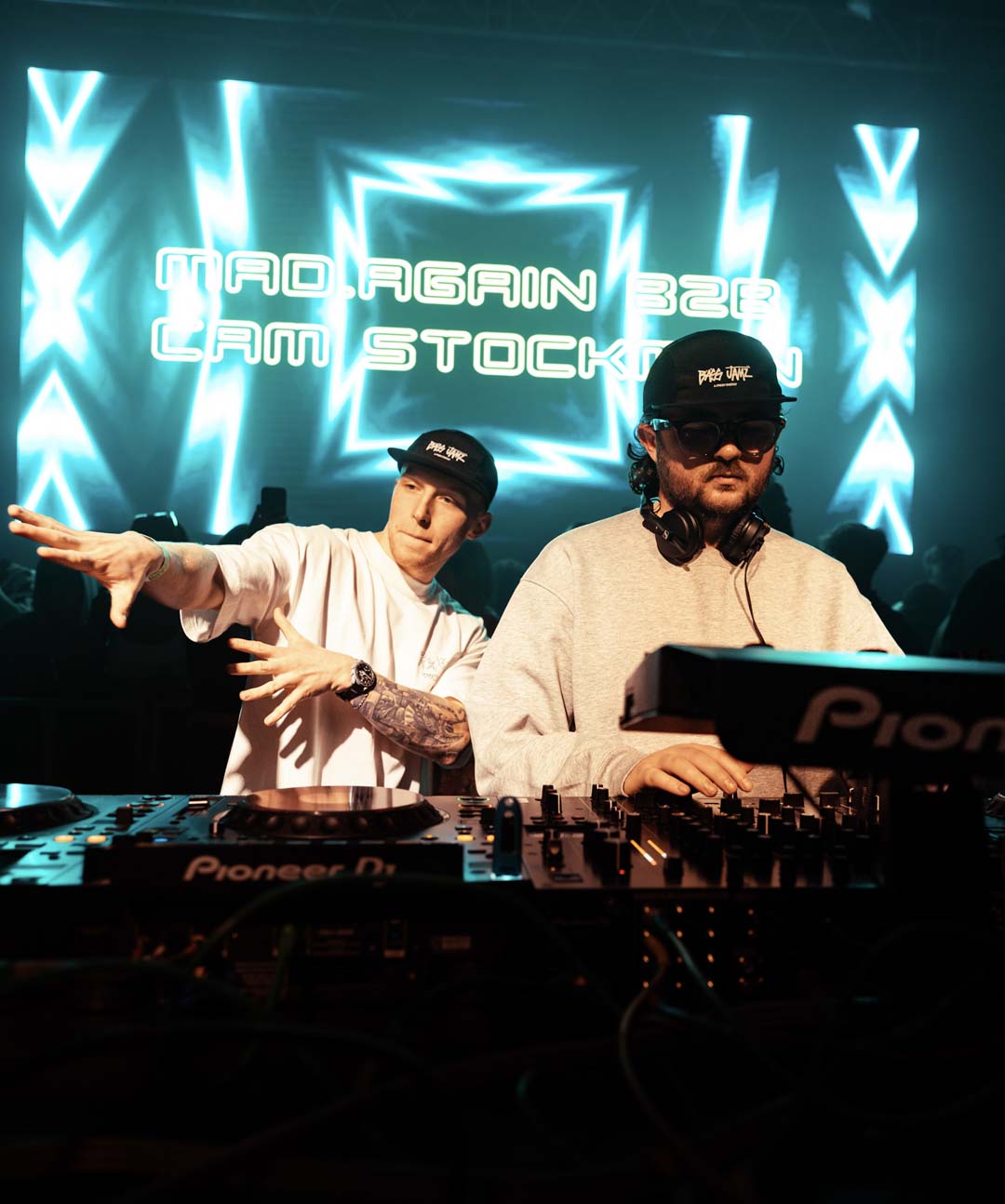Mad Again and Cam Stockman DJs performing at a music event with 'Pioneer' equipment and a vibrant light display.