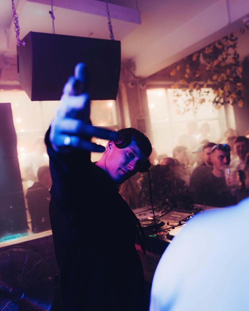 Person taking a selfie in a club setting with DJ equipment and people in the background.