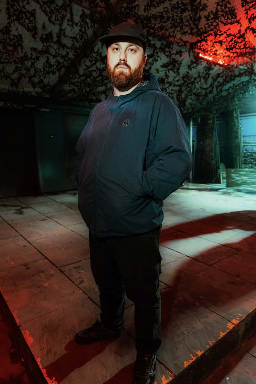 Caul.e wearing a dark jacket and hat standing in a dimly lit area with red lighting.
