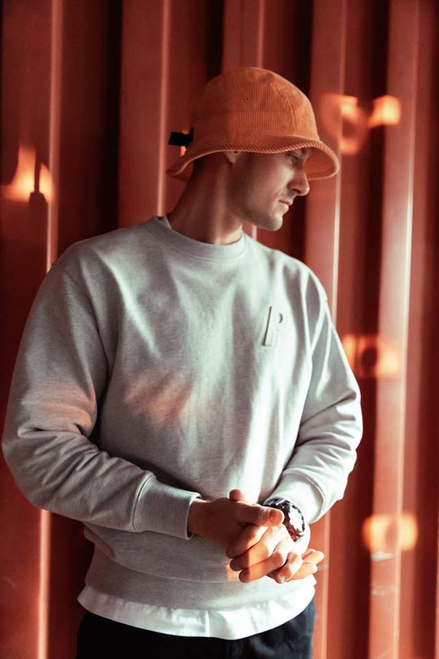 Ellam wearing a light gray sweatshirt with a logo and an orange bucket hat against a warm-toned curtain background.