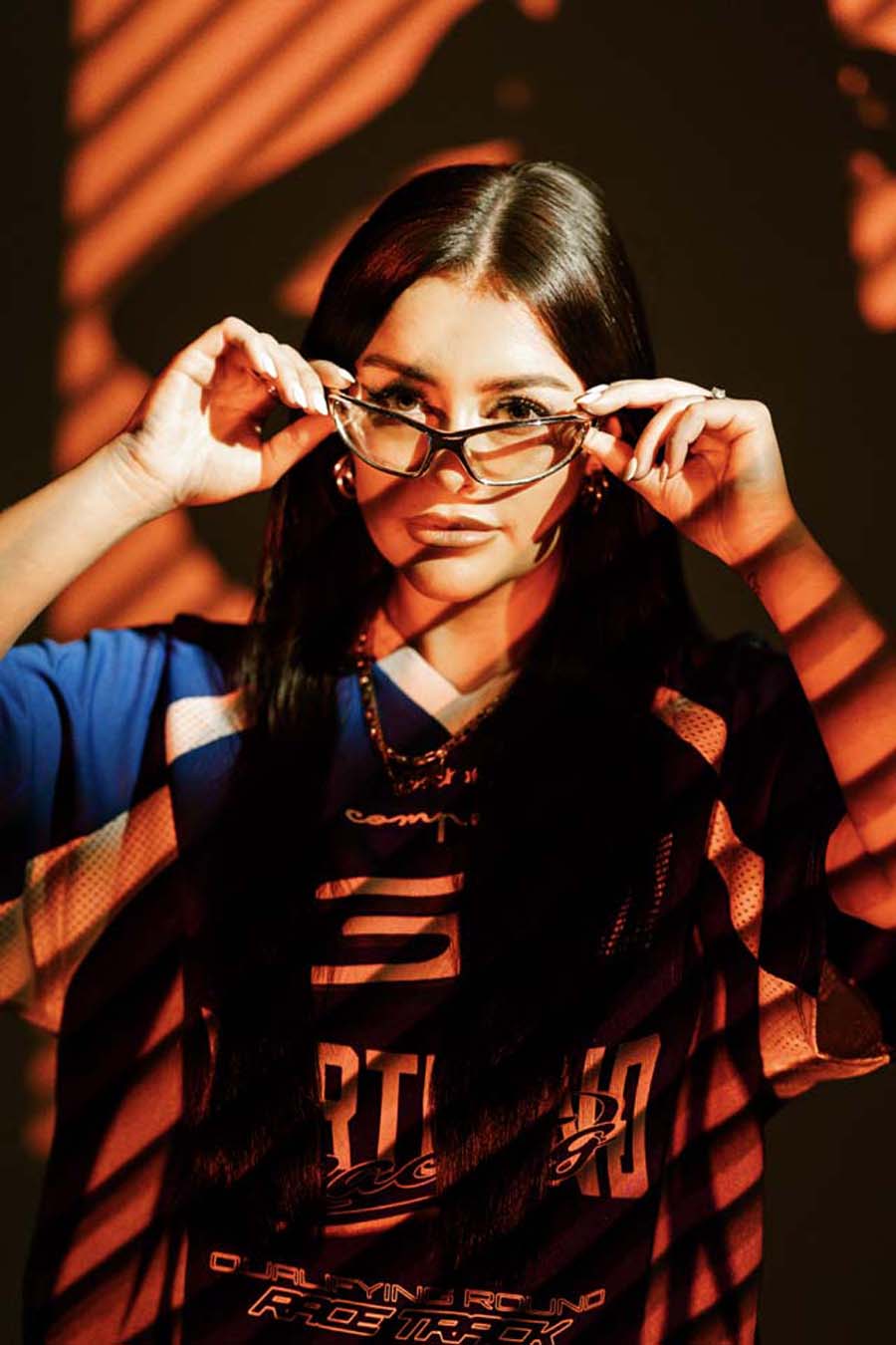 Ellia Jaya wearing a dark jacket with visible branding, adjusting glasses against a warm-toned background.
