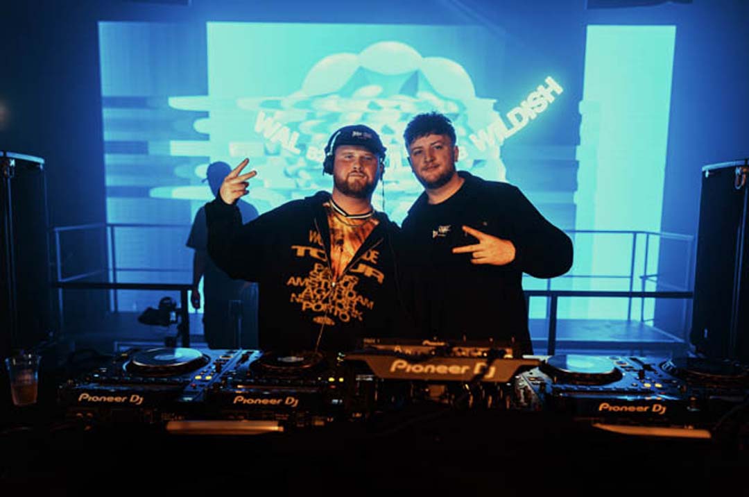 Two DJs posing behind a DJ setup with a colorful screen in the background
