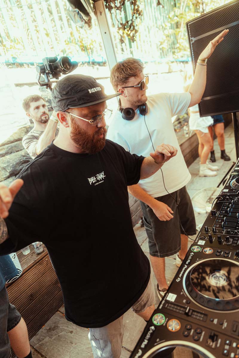 Two DJs at a music event with one operating DJ equipment and the other adjusting settings.