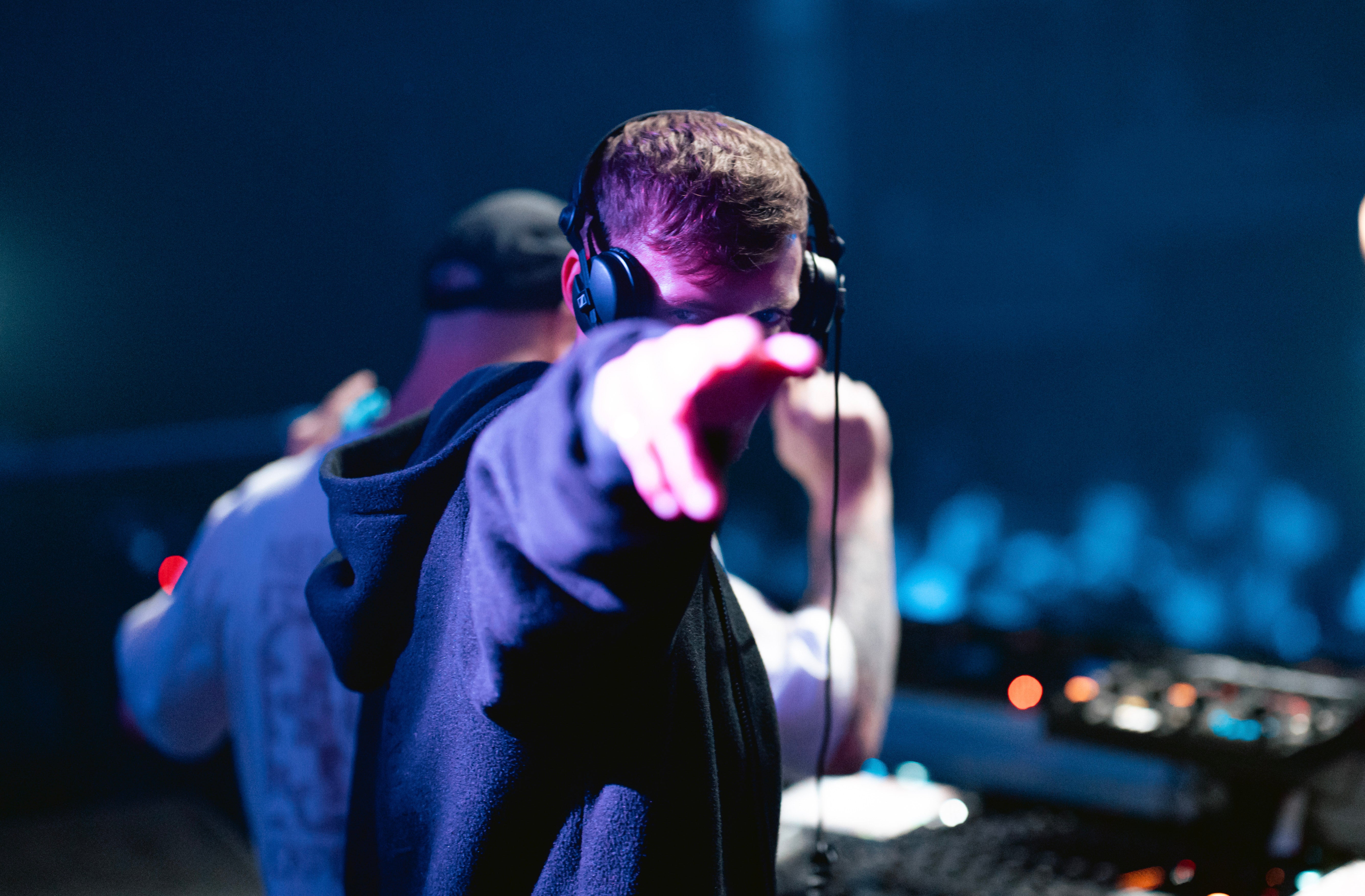 Person wearing headphones pointing forward with a dark, possibly concert or club setting in the background.