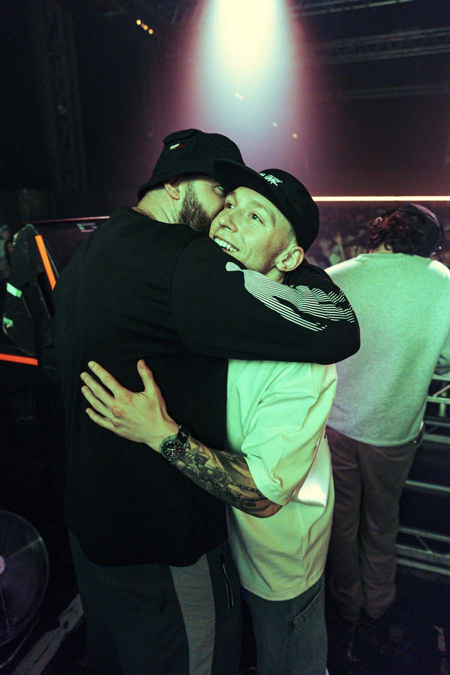 Two dj's hugging on a stage with a spotlight in the background